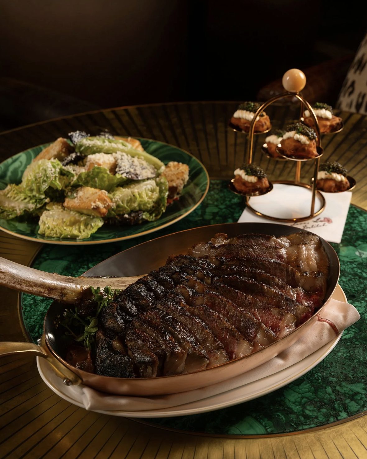 Sliced tomahawk steak in a copper pan beside a Caesar salad and caviar bites on a green malachite table at Delilah