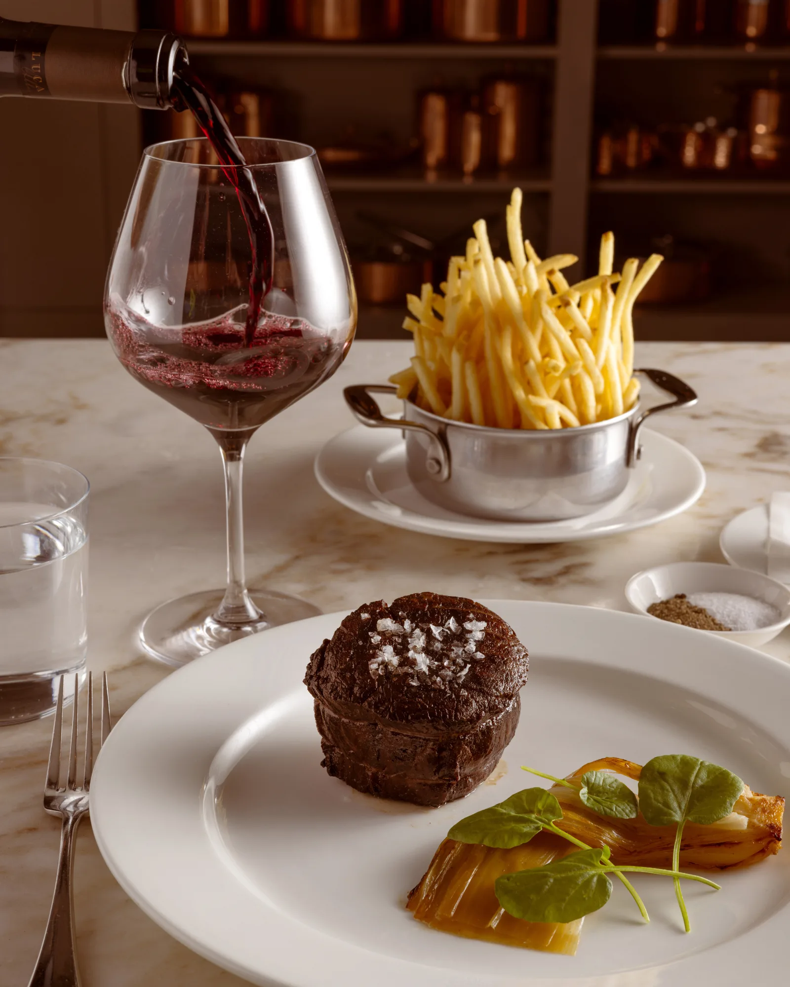 Red wine being poured beside a filet mignon with flaky salt, braised leeks and a pot of fries at The Bedford by Martha Stewart