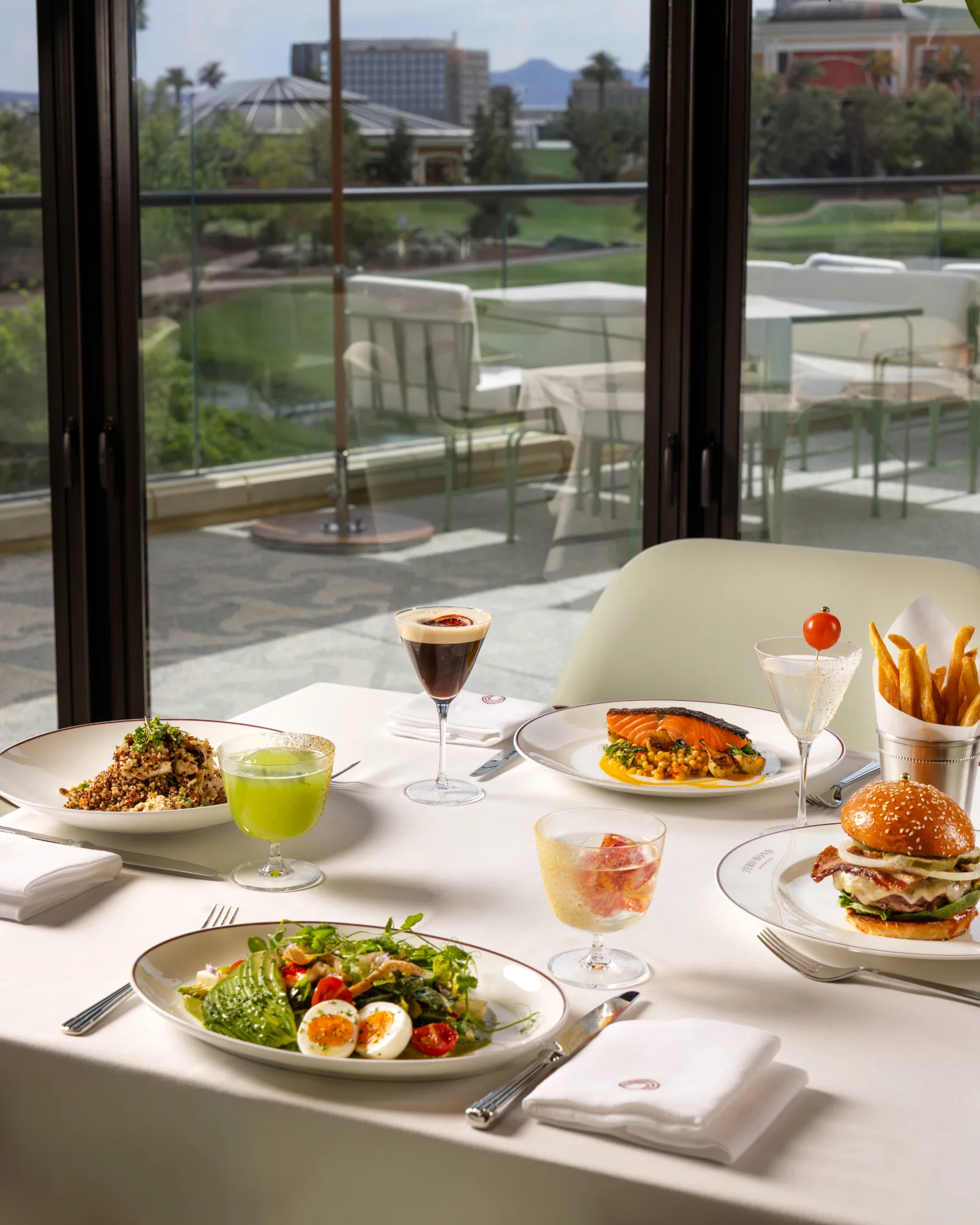 Lunch table at Zero Bond Las Vegas with a burger and fries, salmon, salads and cocktails beside floor-to-ceiling windows